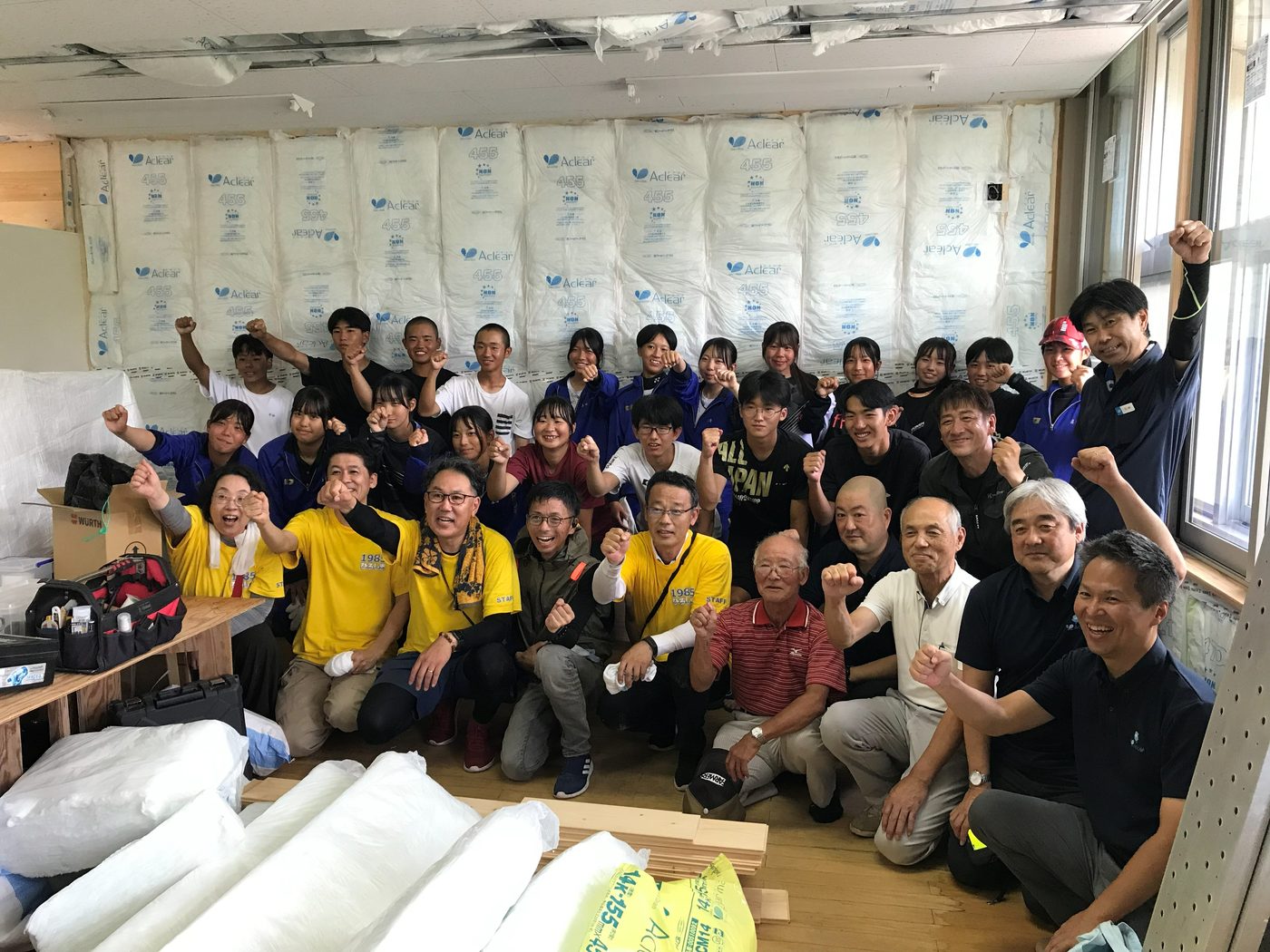 断熱材を貼り終えた壁の前で、参加した中学生と地域の住民、施工スタッフ約40名が並んで拳を上げている集合写真。